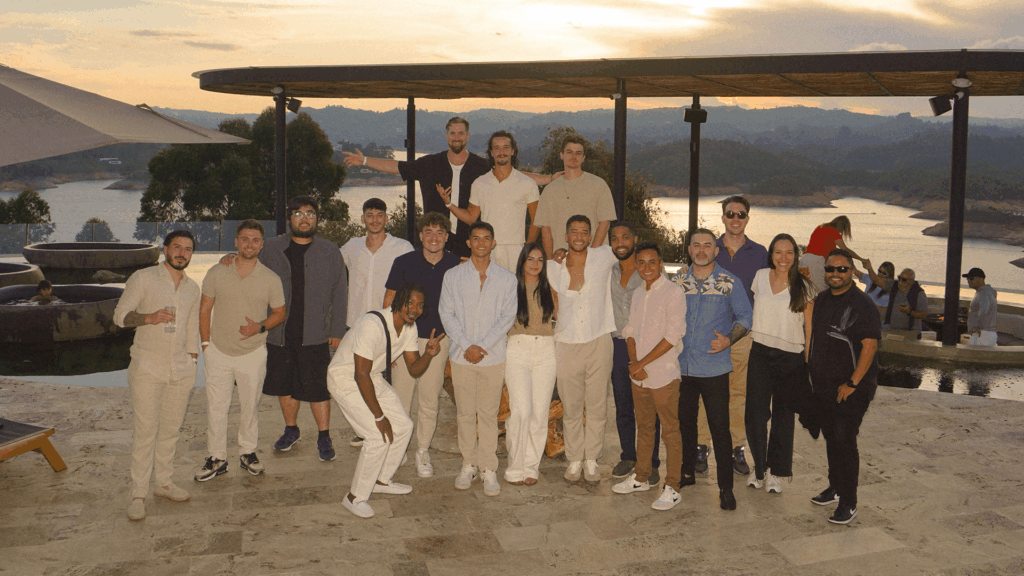 Conquer Mastermind A group of people pose together outdoors at sunset in front of a scenic landscape with water and hills, celebrating their achievements at the Video Agency Mastermind. Video Agency Owner Mastermind