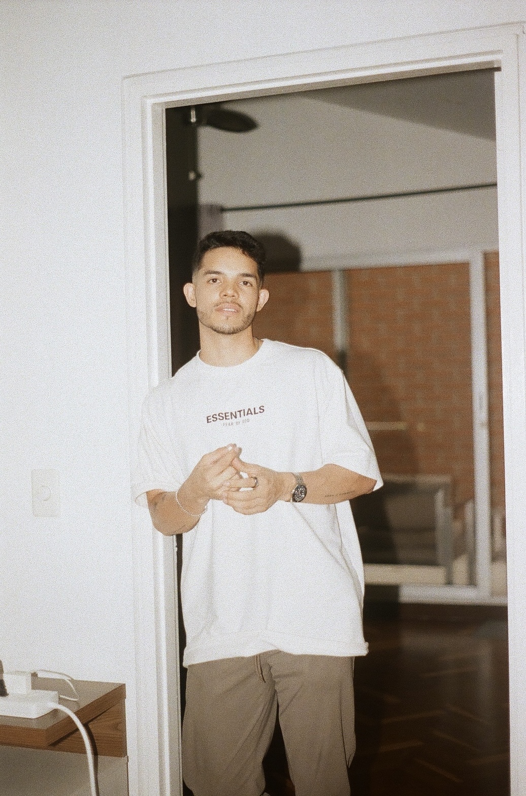 Conquer Mastermind A man stands in a doorway indoors, wearing a white "ESSENTIALS" t-shirt and brown pants, with his hands loosely clasped, embodying the calm confidence often seen at Conquer Mastermind marketing events. Video Agency Owner Mastermind