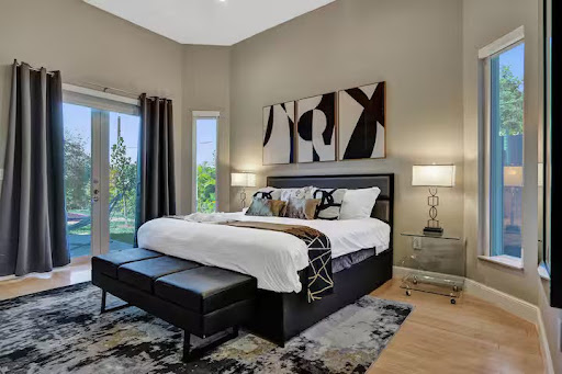 Conquer Mastermind Modern bedroom with a large bed, abstract artwork above the headboard, two windows, glass doors, bedside lamps, and a black bench at the foot of the bed—ideal for relaxing after your latest Conquer Mastermind or Video Agency Mastermind session. Video Agency Owner Mastermind