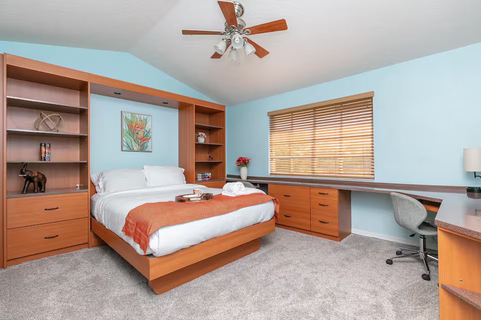 Conquer Mastermind A tidy bedroom with a built-in wood bed and shelving unit, light blue walls, a ceiling fan, a desk with a chair—perfect for late-night Marketing brainstorms—and a window with wooden blinds. Video Agency Owner Mastermind