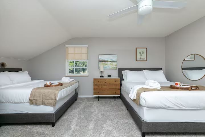 Conquer Mastermind A bedroom with two made double beds, a ceiling fan, a small window, a dresser between the beds, and minimalist decor on light gray walls—perfect for relaxing after a day of Marketing or attending the Conquer Mastermind. Video Agency Owner Mastermind