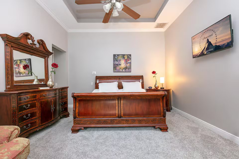 Conquer Mastermind A bedroom with a wooden bed frame, matching dresser and mirror, bedside lamps, two wall art pieces, a ceiling fan, and light grey carpeted flooring—perfect for relaxing after a day of Marketing or Video Agency Mastermind sessions. Video Agency Owner Mastermind