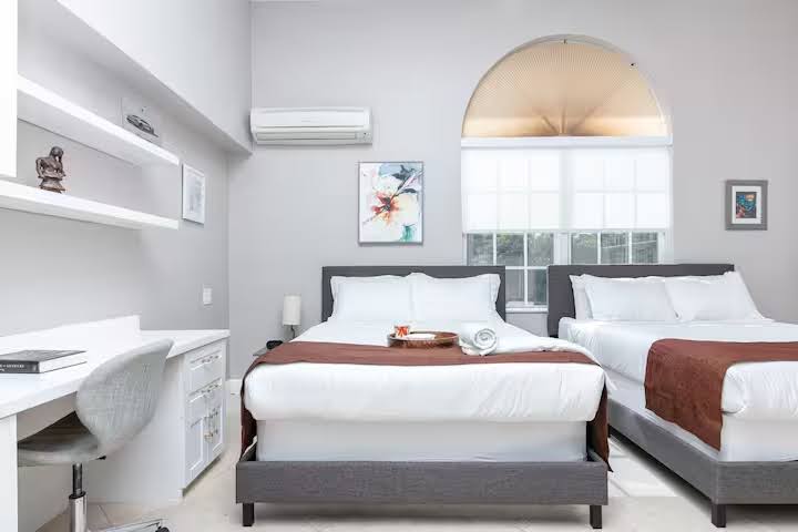 Conquer Mastermind A modern bedroom with two double beds, white bedding, brown throws, a desk for marketing tasks, wall art, shelves, and a large arched window letting in natural light—perfect for relaxing after a Conquer Mastermind session. Video Agency Owner Mastermind