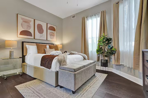 Conquer Mastermind A modern bedroom with a neatly made bed, beige and brown decor, abstract wall art, a potted plant, and natural light from large windows with curtains creates the perfect space to relax after a busy day at the Marketing or Conquer Mastermind. Video Agency Owner Mastermind