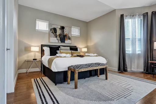 Conquer Mastermind Modern bedroom with a large bed, abstract art above the headboard, two bedside tables with lamps, and a sleek wooden bench—perfect for relaxing after a long day at your latest Marketing or Conquer Mastermind event. Video Agency Owner Mastermind