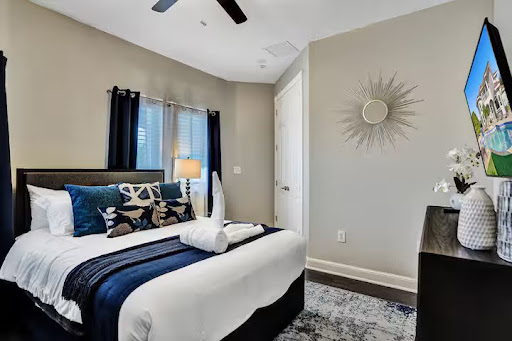 Conquer Mastermind Modern bedroom with a neatly made bed, navy and white bedding, decorative pillows, a lamp by the window, wall-mounted TV—perfect for relaxing after a busy day of Marketing or brainstorming with your Conquer Mastermind group. Video Agency Owner Mastermind