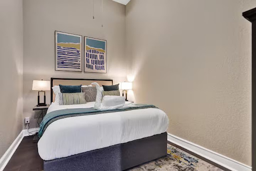 Conquer Mastermind A neatly made bed with pillows and folded towels, flanked by two lamps, sits against a beige wall with two abstract paintings above the headboard—perfect for relaxing after a productive Conquer Mastermind session. Video Agency Owner Mastermind
