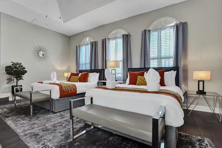 Conquer Mastermind Modern bedroom with two double beds, red and white bedding, three arched windows with gray curtains—perfect for relaxing after a Conquer Mastermind or Marketing brainstorming session. Features two benches, lamps, a plant, and sunburst wall decor. Video Agency Owner Mastermind