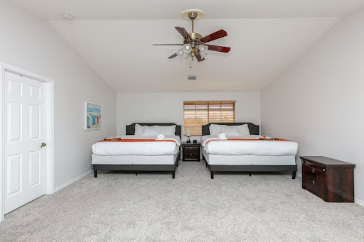 Conquer Mastermind Two neatly made double beds with white bedding and orange runners in a spacious, carpeted bedroom—perfect for relaxing after an inspiring day at a Video Agency Mastermind or Conquer Mastermind marketing event. Ceiling fan and window with blinds included. Video Agency Owner Mastermind