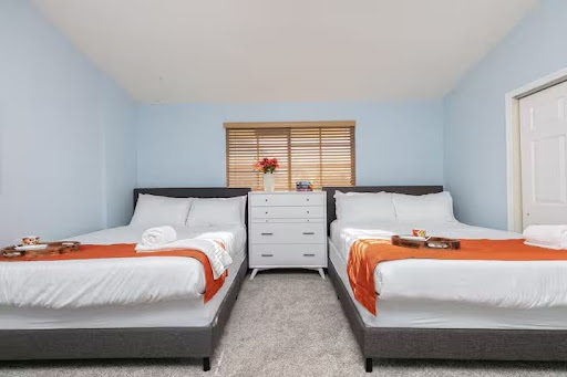 Conquer Mastermind A bedroom with two double beds, each with white linens and orange throws, a white chest of drawers between them, and a window with wooden blinds above—perfect for unwinding after a day at the Conquer Mastermind marketing event. Video Agency Owner Mastermind